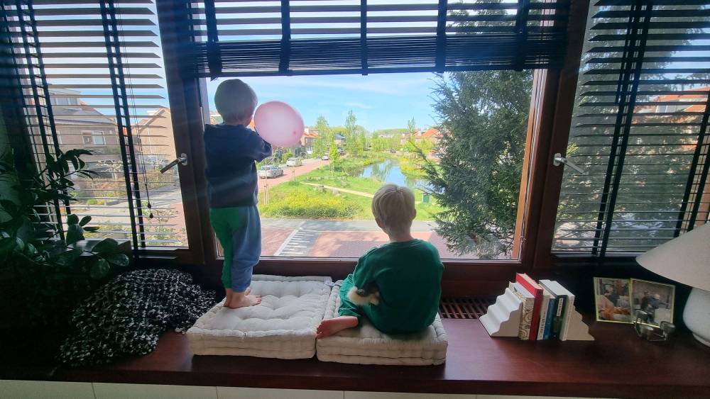 Two children stand on a windowsill looking out at a canal view, one holding a pink balloon.