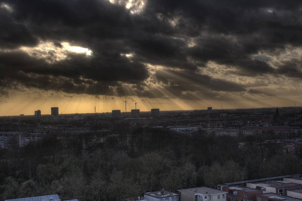 Sunbeams break through heavy dark clouds over a city skyline.