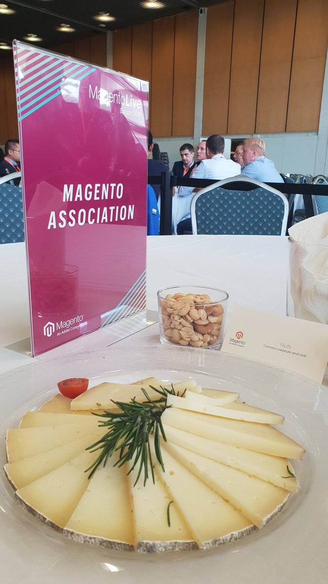 A sliced cheese platter sits on a table beside a Magento Association standee.