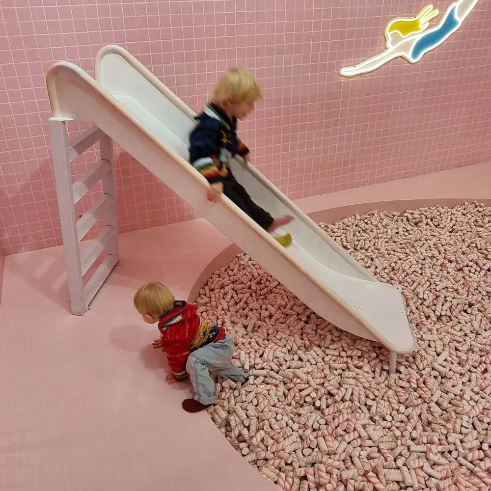 Two children going down a slide into a pit of light-colored balls in a pink-tiled play area.