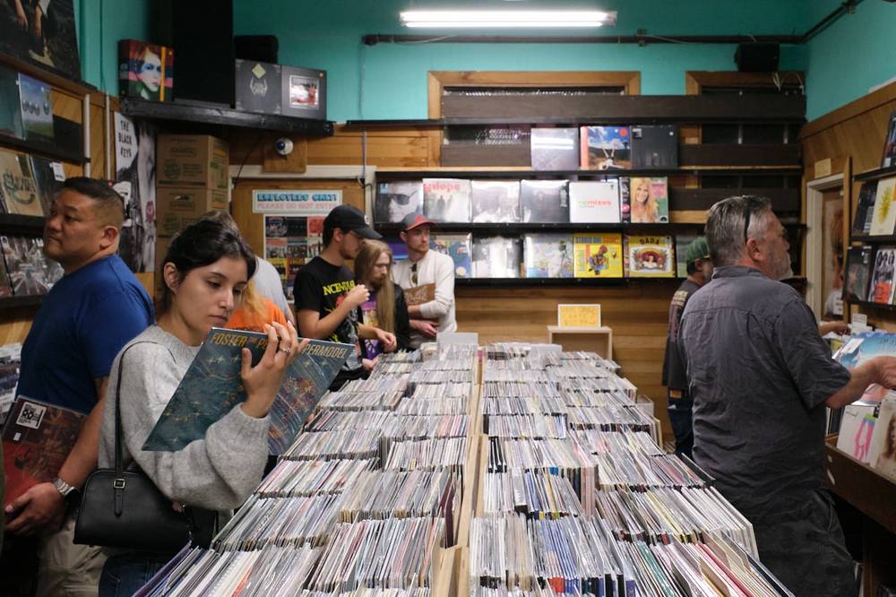 Customers browse long center bins of vinyl records in a narrow aisle at Music Millennium. In the foreground, a woman in a gray sweater flips through records while holding up a Foster the People "Supermodel" LP. Shelves of featured album covers line the teal-painted back wall beneath a fluorescent light.