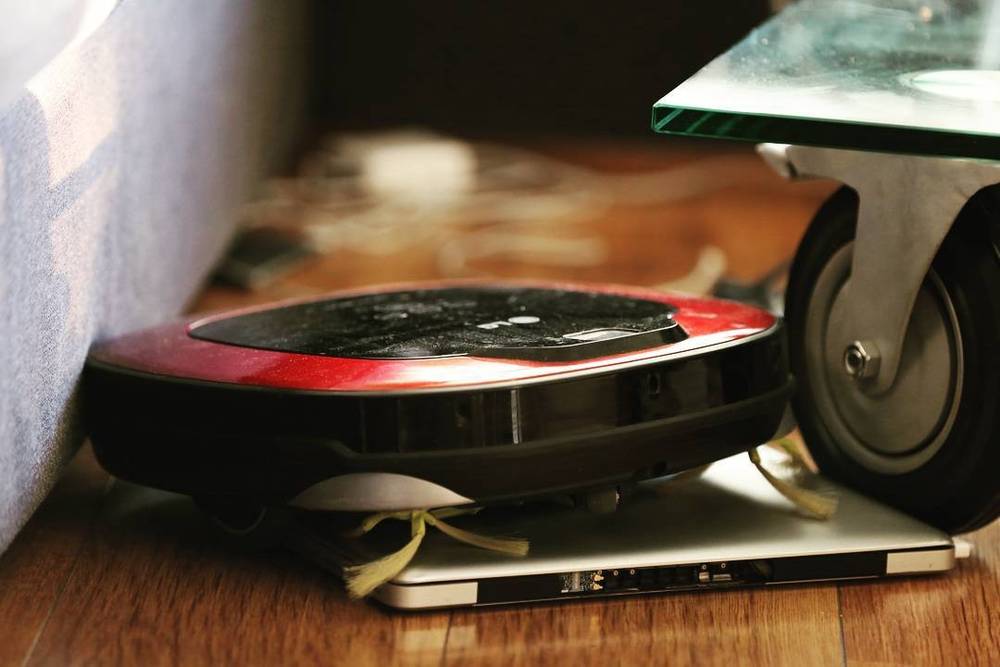 Close-up of a robot vacuum cleaner wedged against a laptop on a wooden floor.