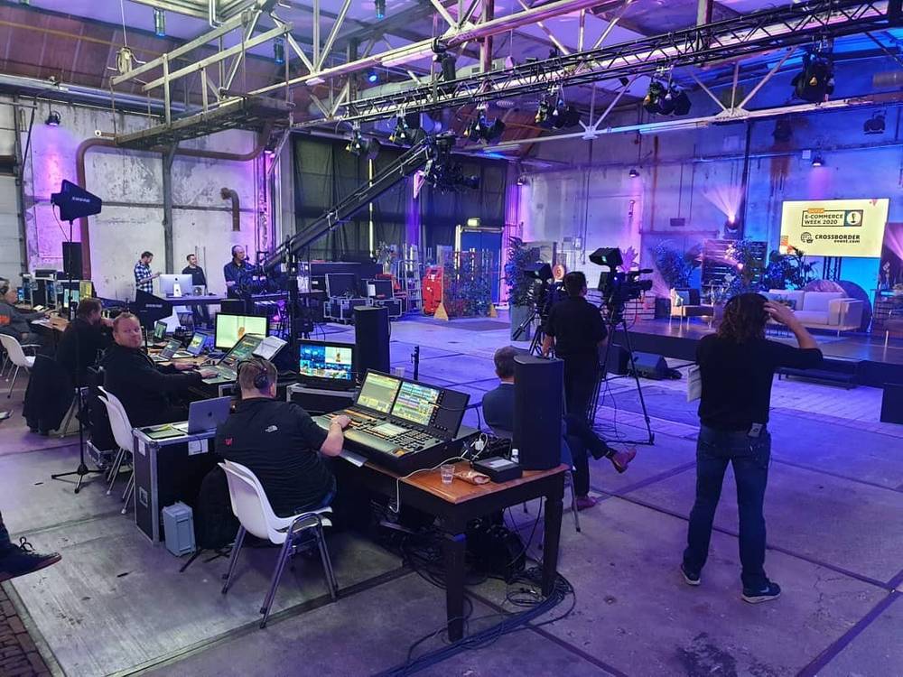 A television production crew working at mixing desks and camera equipment inside an industrial venue set up for a livestreamed conference.