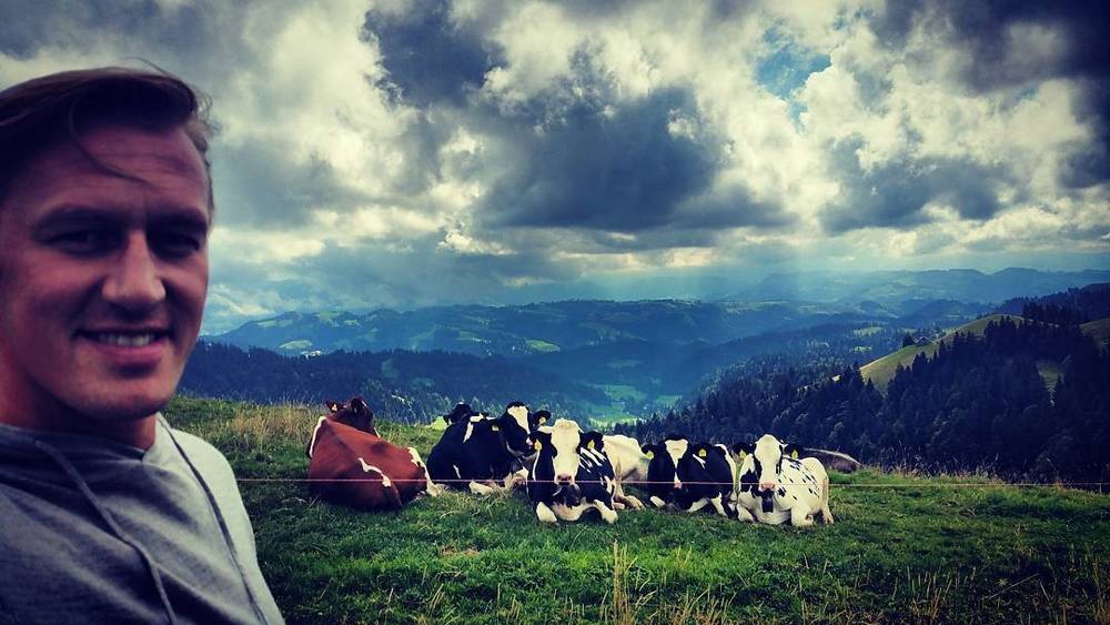 A selfie of a smiling man on a hillside overlooking dramatic clouds, with cows resting in the grass beside him.