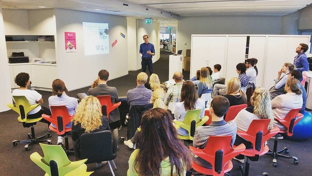 An office presentation with a speaker addressing an audience seated on orange and green chairs.