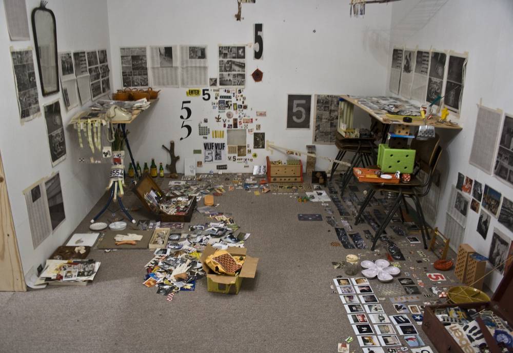 A cluttered art installation room with photos, papers and objects scattered across the floor and walls.
