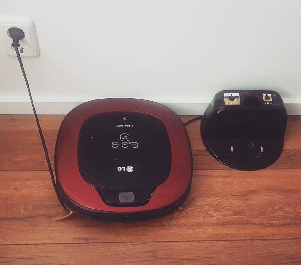 A red robot vacuum cleaner parked next to its charging dock on a wooden floor.