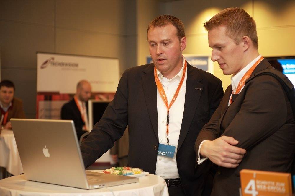 Two men in suits with conference lanyards looking closely at a laptop screen at an expo booth.