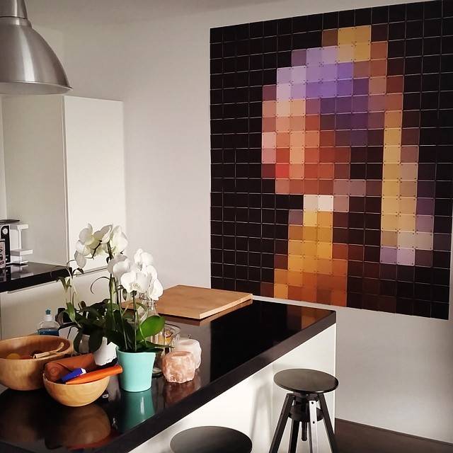 A pixelated wall artwork resembling Vermeer's 'Girl with a Pearl Earring' hangs in a modern kitchen with flowers on the counter.