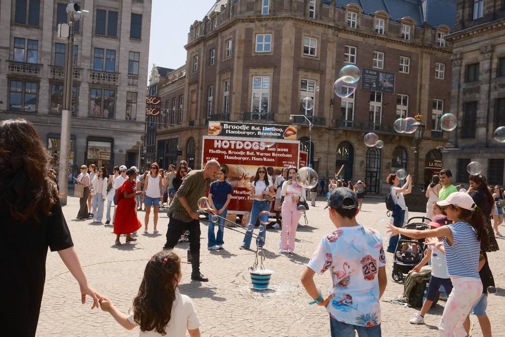 Mother and daughter hold hands while watching a man blow comically large bubbles in Dam Square