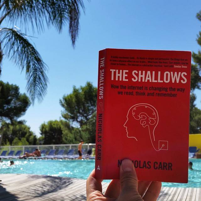 A hand holds up the book 'The Shallows' by Nicholas Carr beside a swimming pool with palm trees.