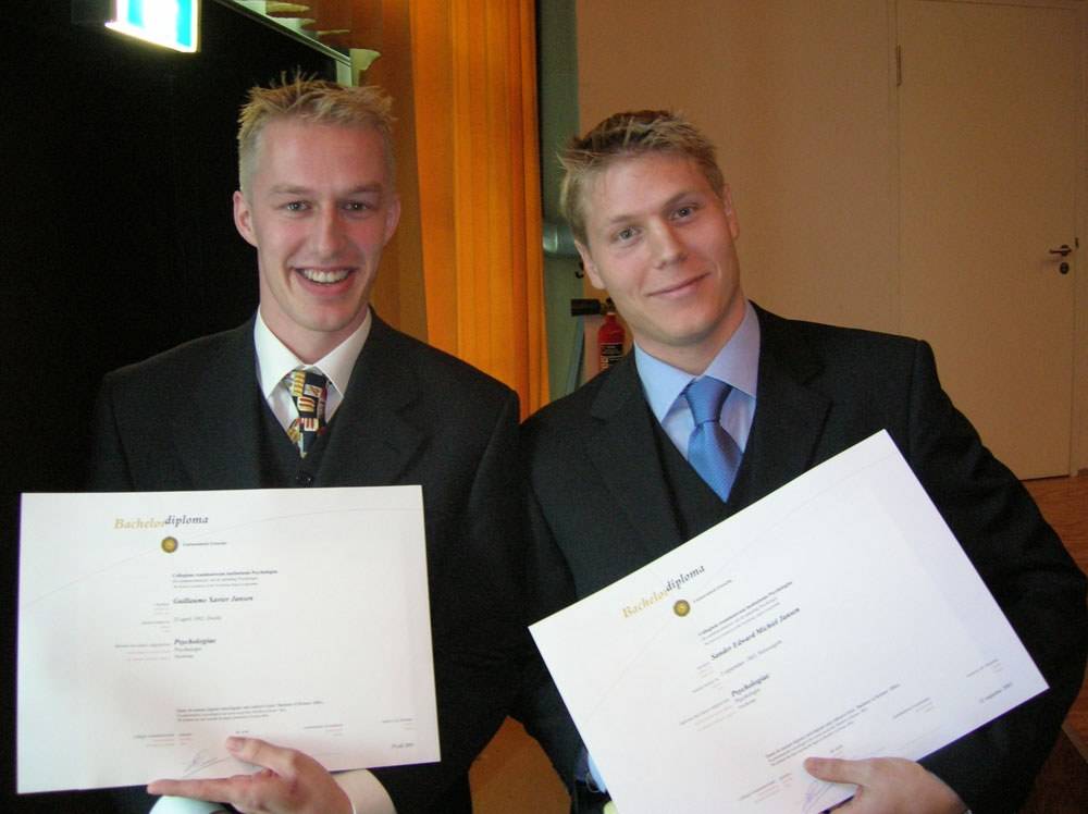 Two young men in suits smile while holding their bachelor's diplomas.