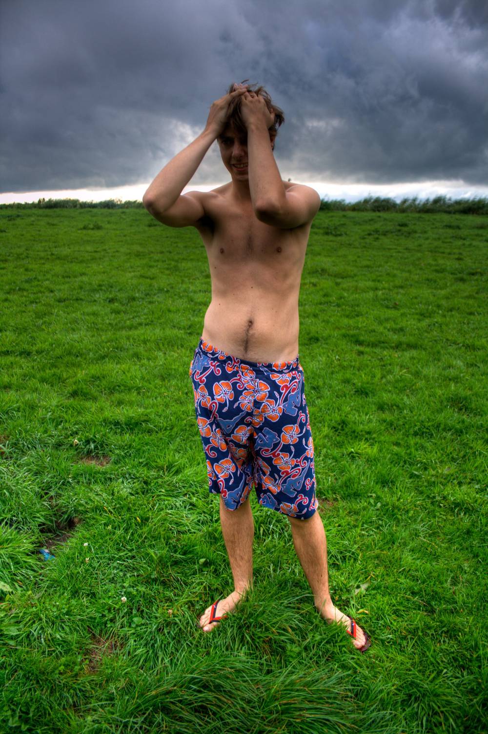 A shirtless man in swim shorts stands in a grassy field with his hands clasped over his head under a stormy sky.
