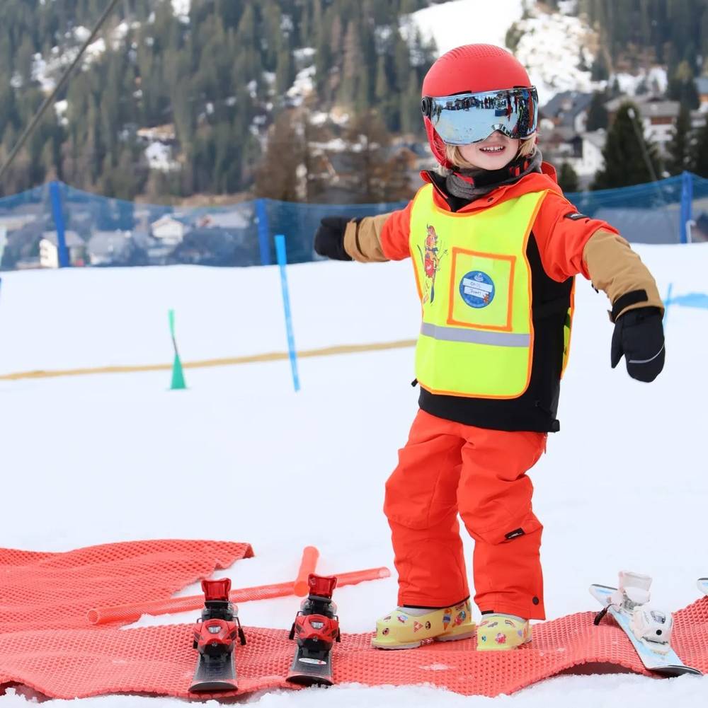 A child in full ski gear and goggles smiling on a beginner slope, with small skis and boots laid out nearby.