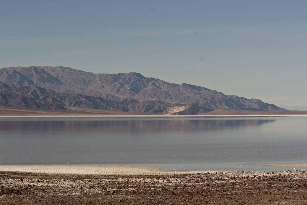Layered desert mountains are reflected in the still water of a shallow salt lake.