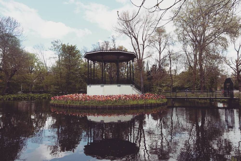 A bandstand pavilion on an island surrounded by a pond in a park, with reflections in the water.