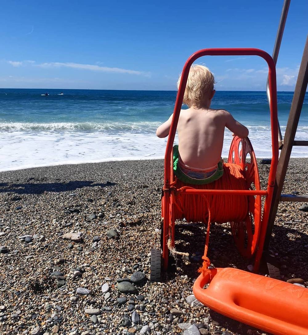 A shirtless child sitting on a red lifeguard chair on a pebble beach, facing the sea.