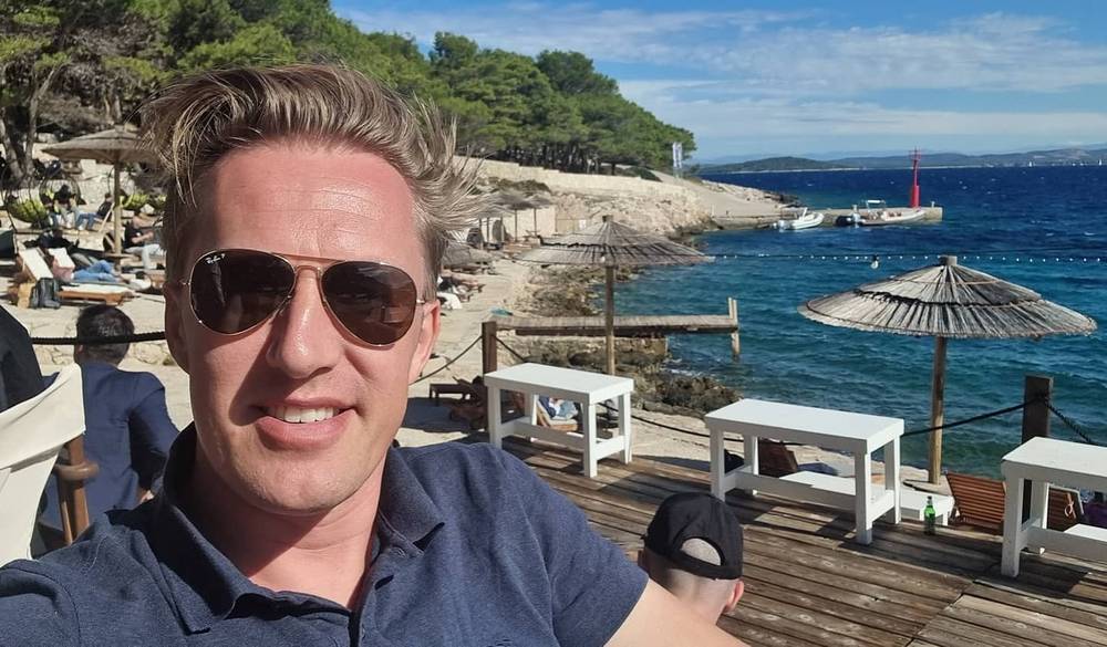 Guido smiling in sunglasses on a wooden beach deck with sun loungers and thatched umbrellas by the sea.
