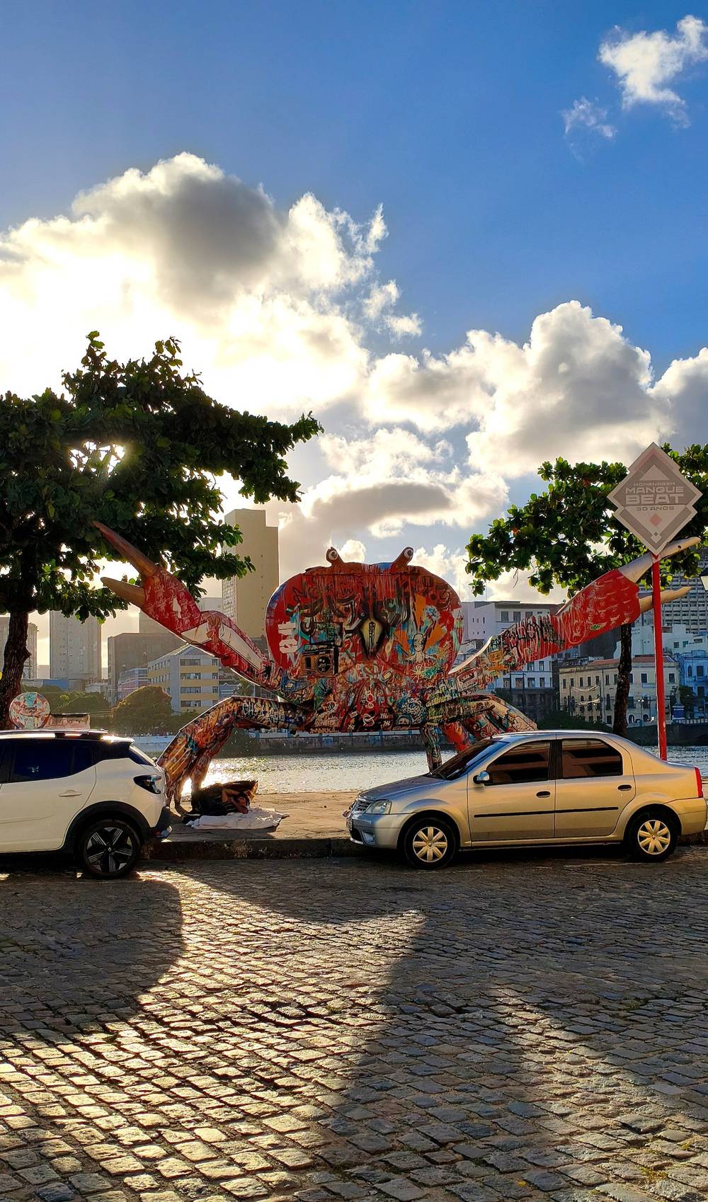 Foto colorida de uma grande escultura de caranguejo vermelho coberto de graffiti, numa calçada,  em frente a escultura carros estacionados ao fundo o rio, a cidade e o céu azul com nuvens ilinadas pelo sol do fim de tarde