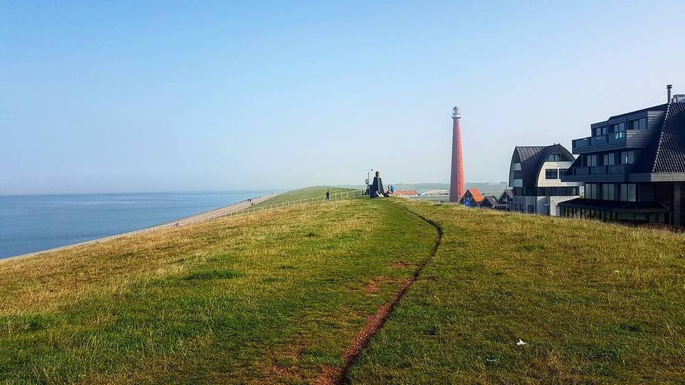 A grassy dune path leading toward a red lighthouse and houses along the coast.