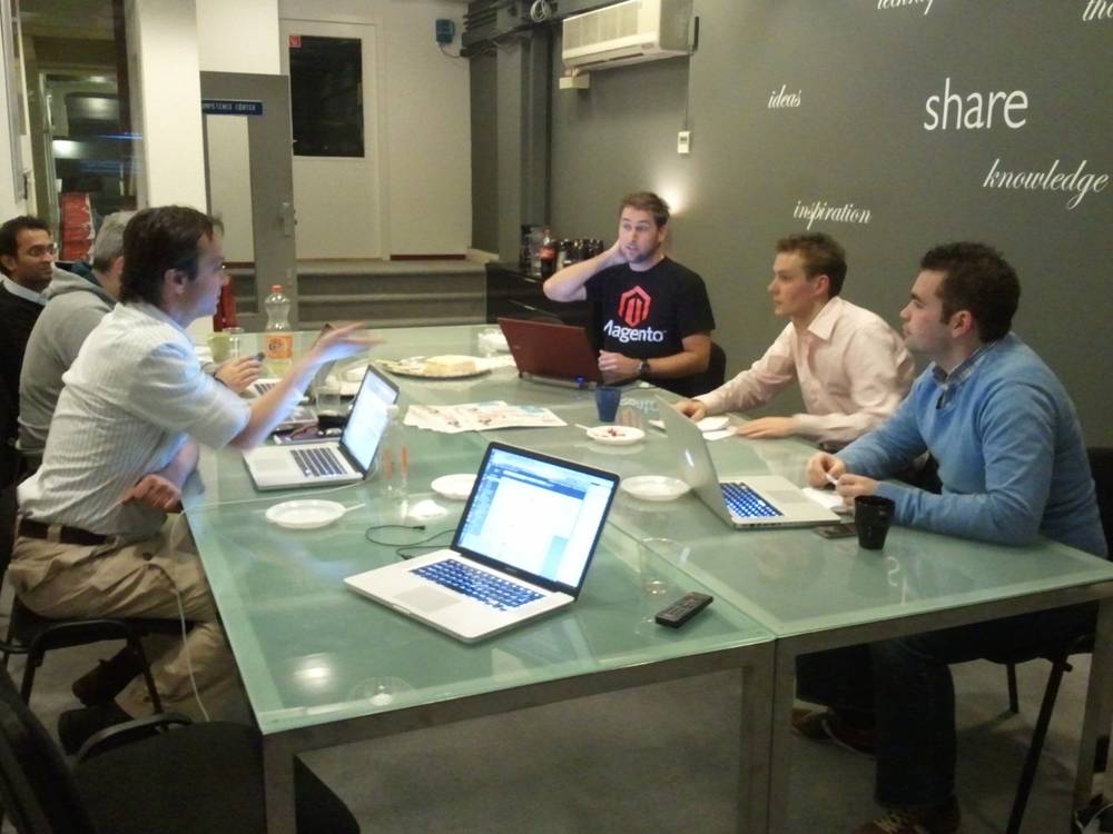 Four colleagues gather around a glass table with laptops, one wearing a Magento t-shirt, deep in discussion.