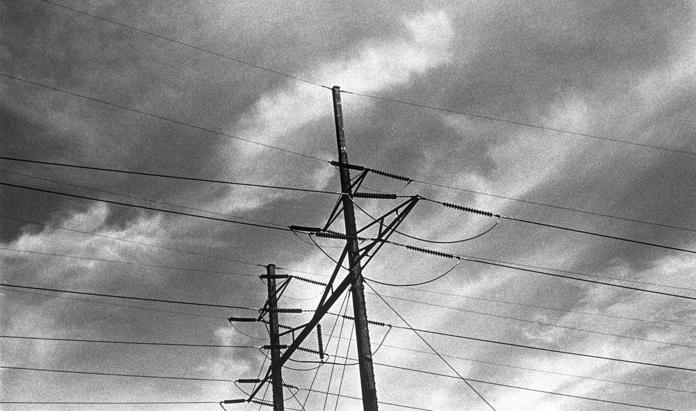 Two transmission poles with crossarms and ceramic insulators rise against a turbulent sky of streaked cumulus clouds. Power lines radiate outward across the frame in multiple directions, drawing taut diagonals over the upper half of the image. The poles, silhouetted in dark grey, dominate the right side. The left opens onto bright sky.