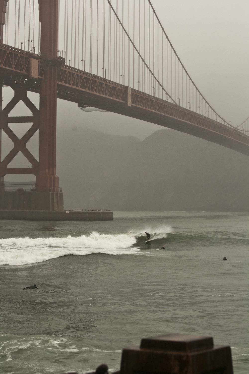 A surfer rides a wave near the bridge tower, seen through misty fog.