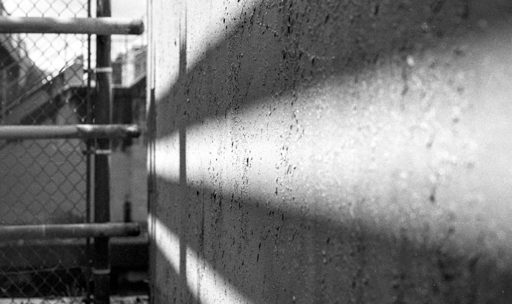 View along a textured concrete wall receding sharply into the distance, its pebbled aggregate surface streaked with vertical drip stains. Two pale horizontal bands of light cross the wall at upper and lower thirds. To the left, a chain-link fence with tubular steel rails runs parallel to the wall, opening onto a blurred industrial background of low buildings and overcast sky.