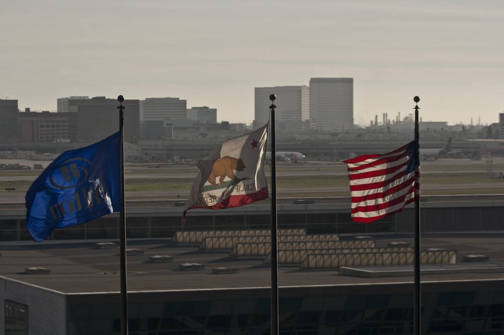 Flags including the US and California state flag fly in front of an airport terminal and skyline.