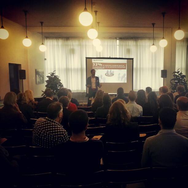 A speaker addresses an audience in front of a screen showing a statistic, in a room with hanging globe lights.