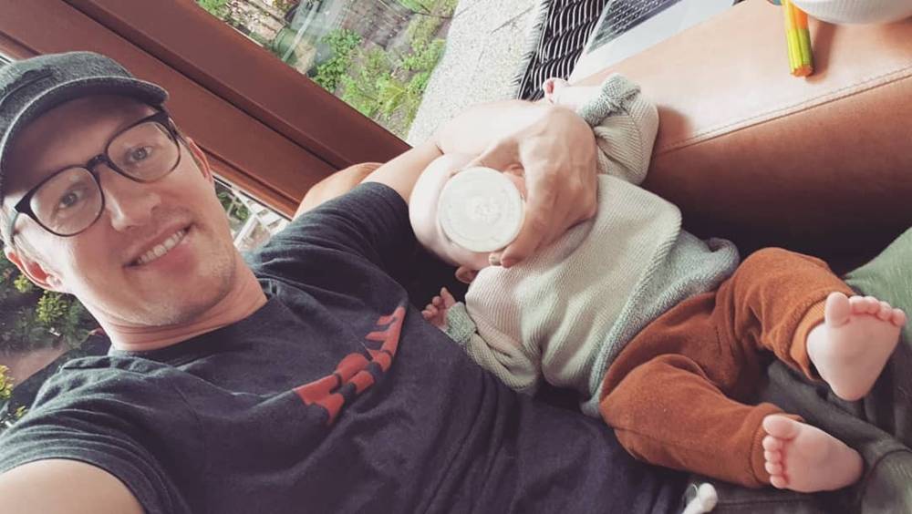 A man feeds a baby a bottle while smiling outdoors on a balcony.