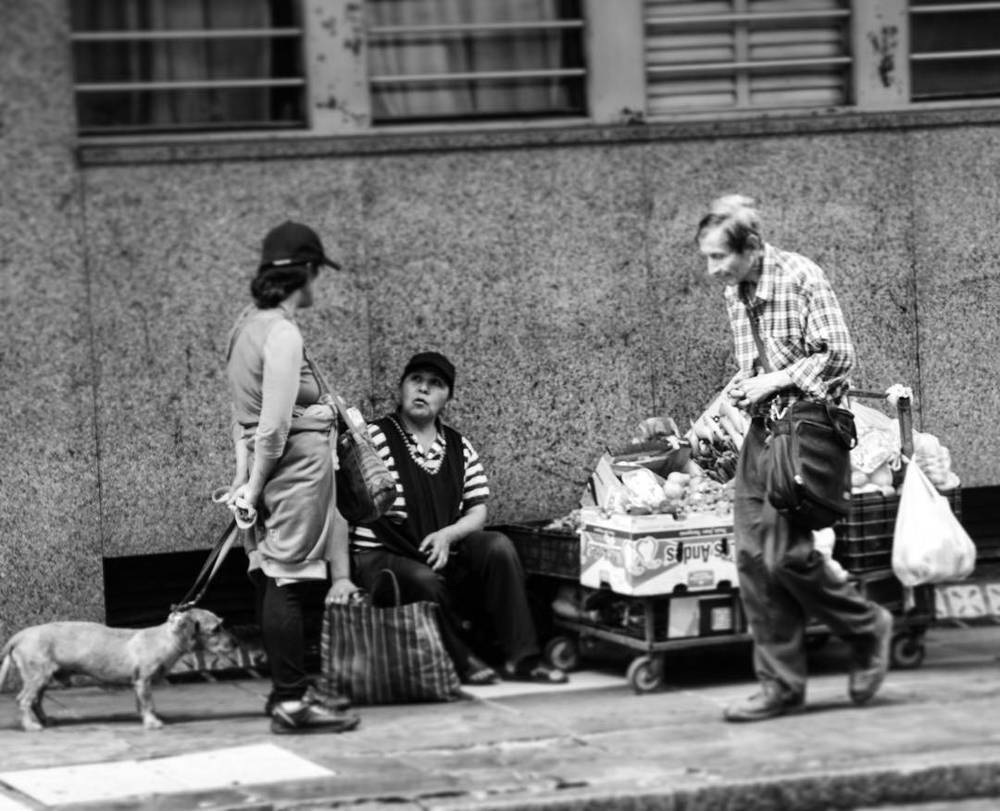 A black and white photo of three people and a small dog resting on the sidewalk beside carts of belongings.
