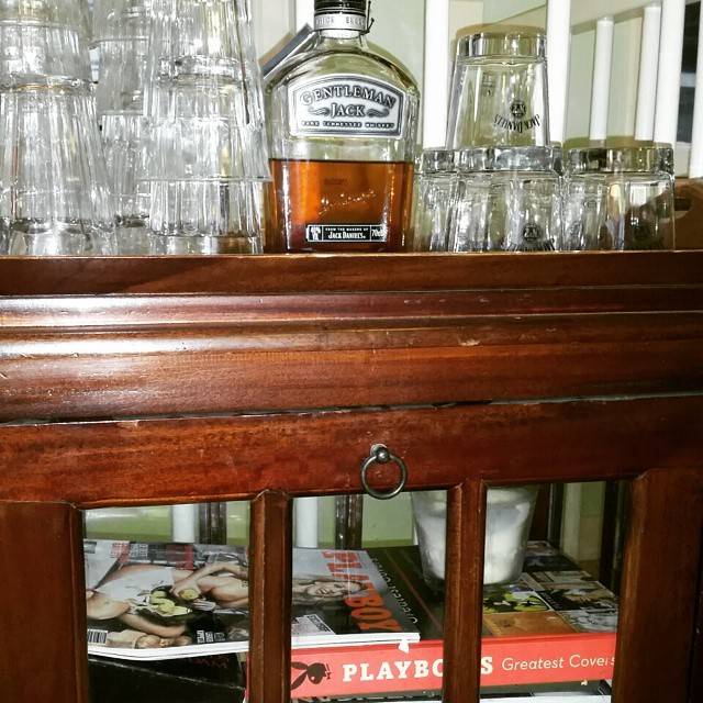 Bottles of whiskey and glasses displayed on a wooden bar shelf, with magazines below.