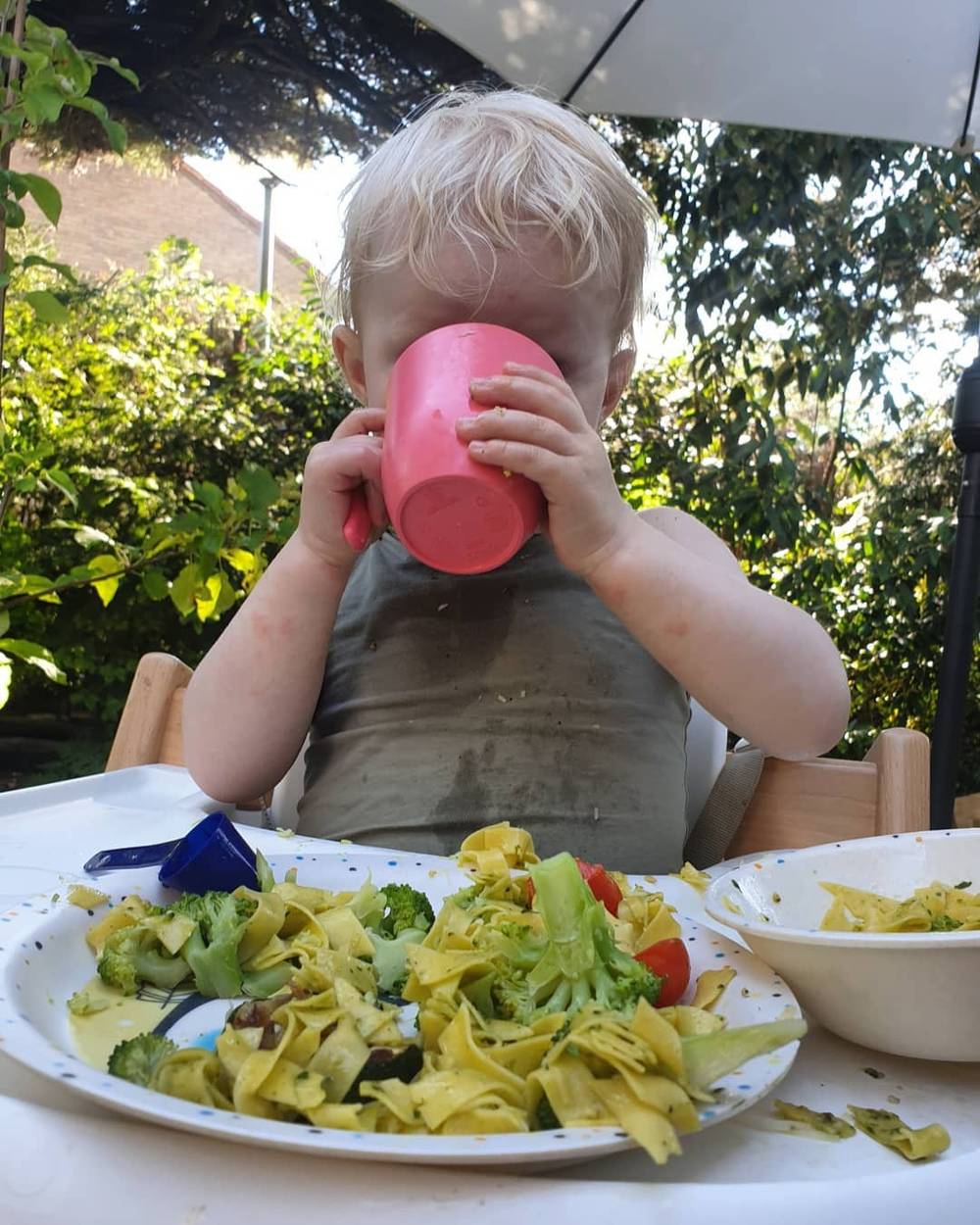 A toddler drinking from a pink cup at an outdoor table, with a plate of pasta and vegetables in front of him.