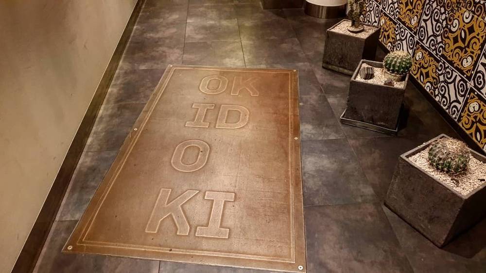 A floor mat spelling "OK ID O KI" sits at a hotel entrance flanked by potted cacti.