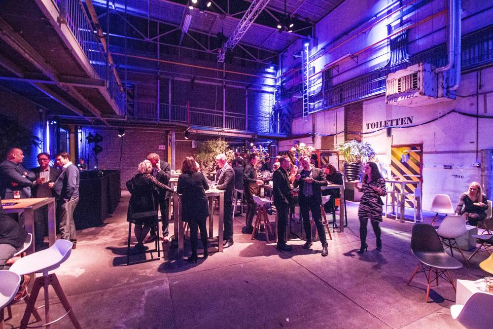 Guests sit and chat at high tables near a 'Toiletten' sign in the industrial hall.