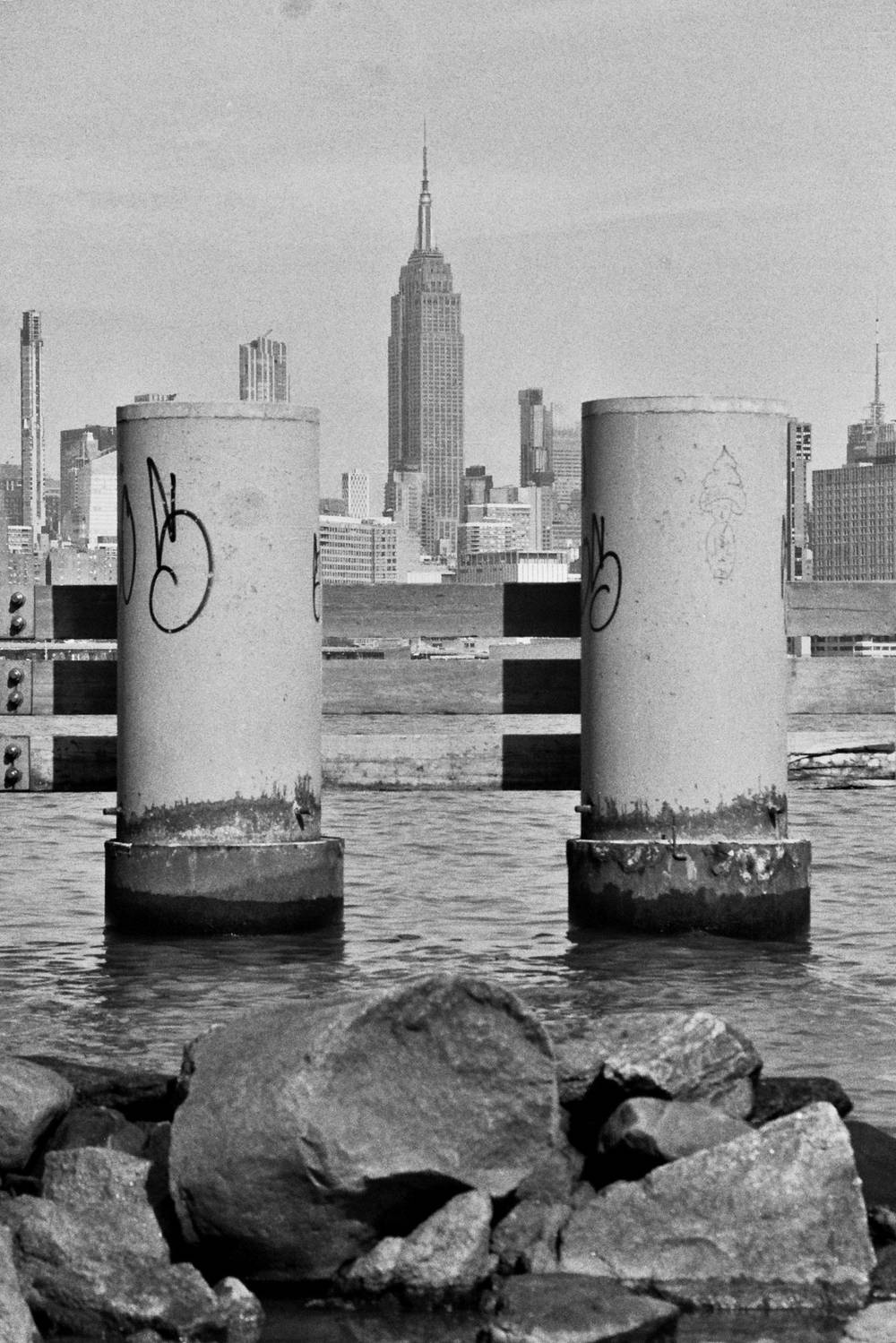 View of the Empire State Building from across the East river, framed by two concrete pillars near the shore. Black and white.