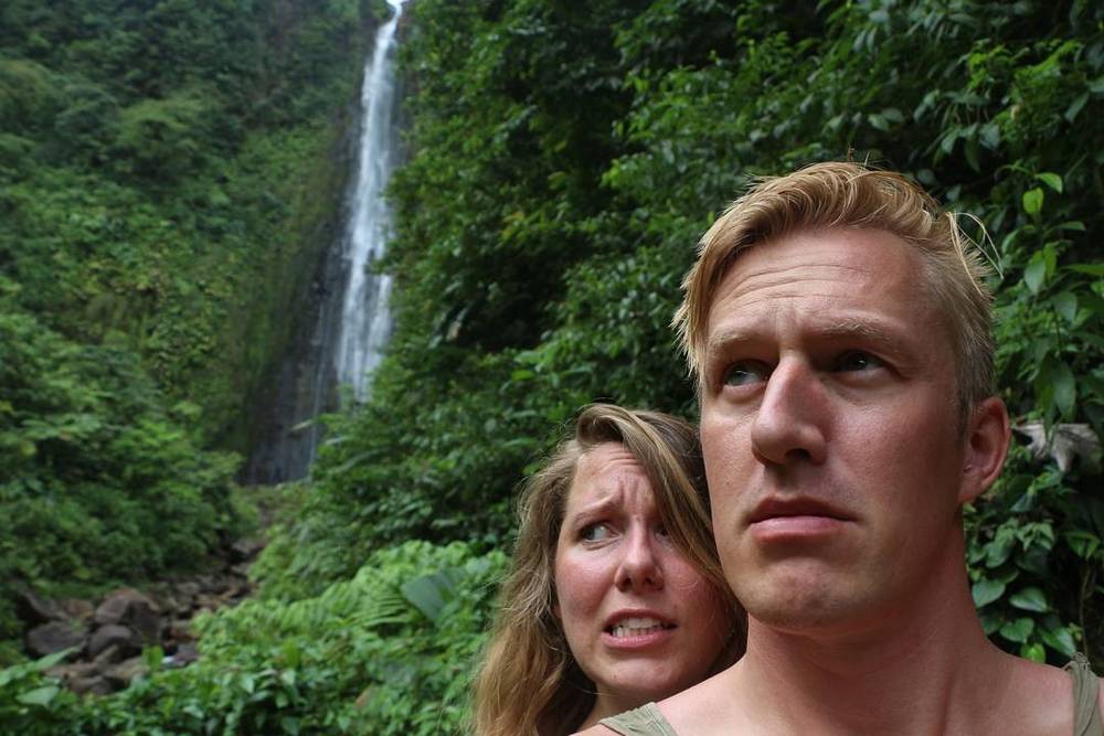 A couple making puzzled faces in front of a jungle waterfall.