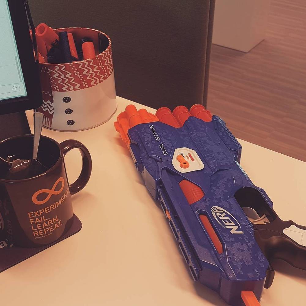A desk with a mug reading "Experiment, Fail, Learn, Repeat" next to a foam dart toy gun.