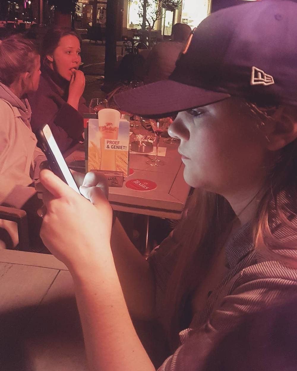 Two women at a café table, one checking her phone while wearing a cap.