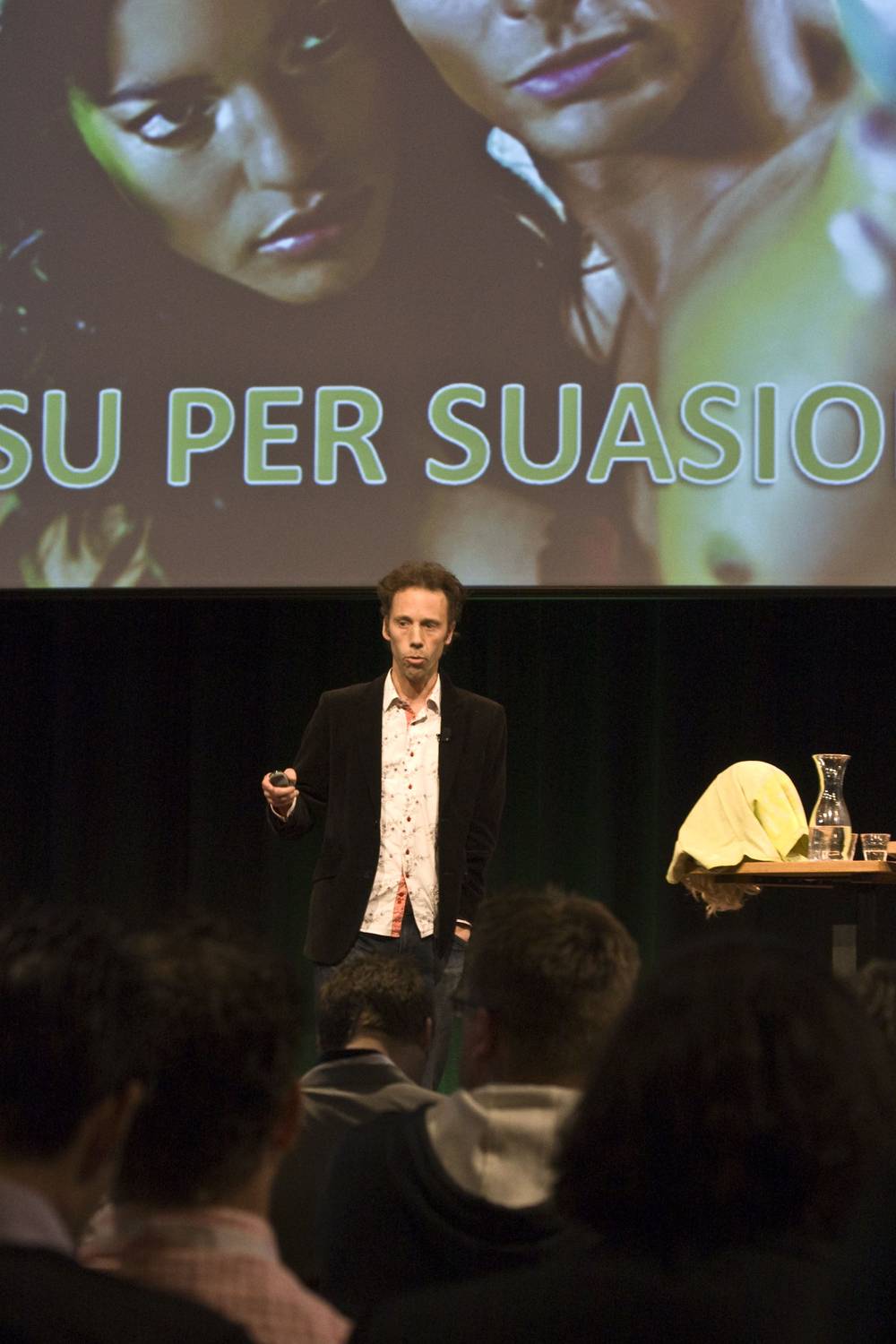 A speaker gestures on stage in front of a screen reading 'SU PER SUASION'.
