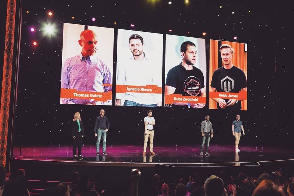 Four men on stage under spotlights, their names displayed above them at a conference.