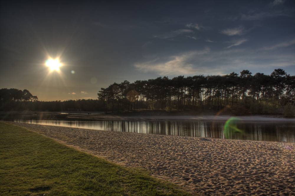 The sun glows low over Henschotermeer lake beyond a sandy beach and treeline.