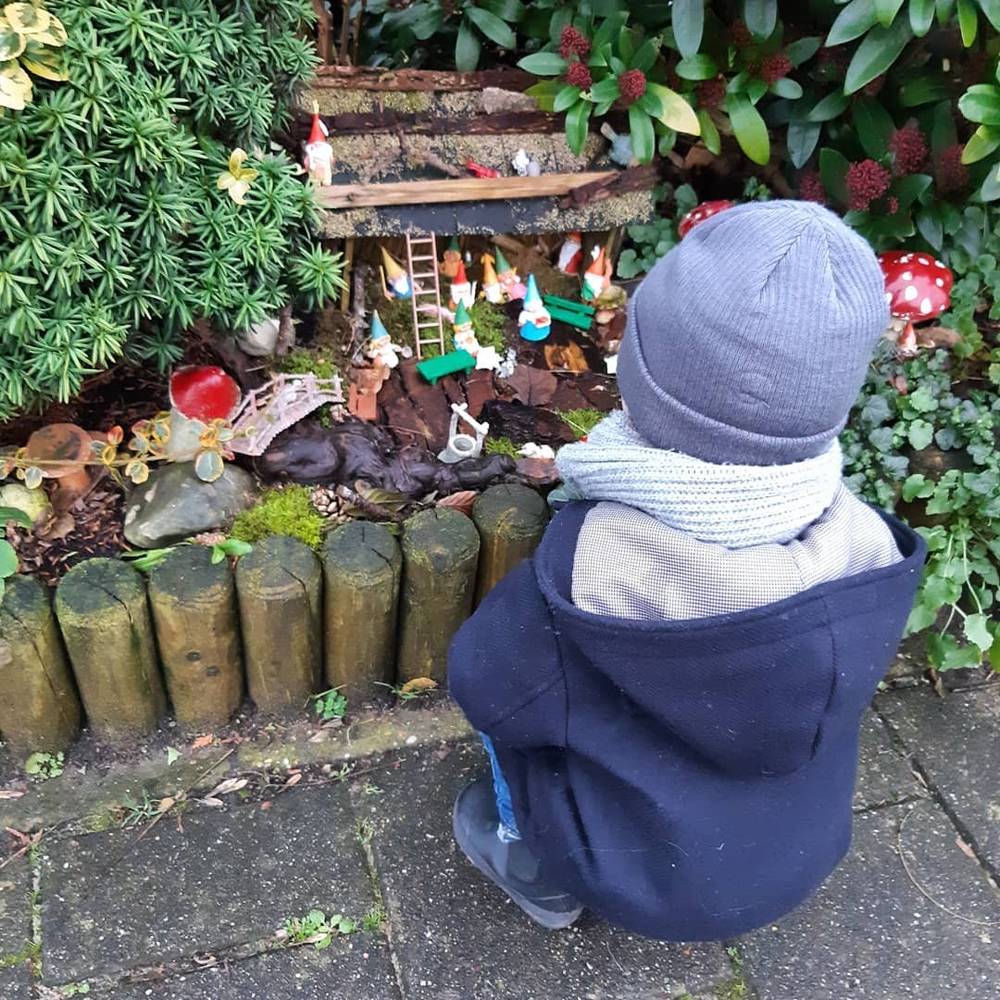 A child in a blue coat looking at a miniature garden gnome village display tucked among plants.