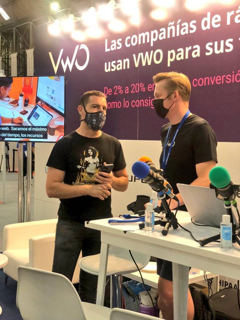 Two men in face masks talking into microphones on a conference stage in Spain.