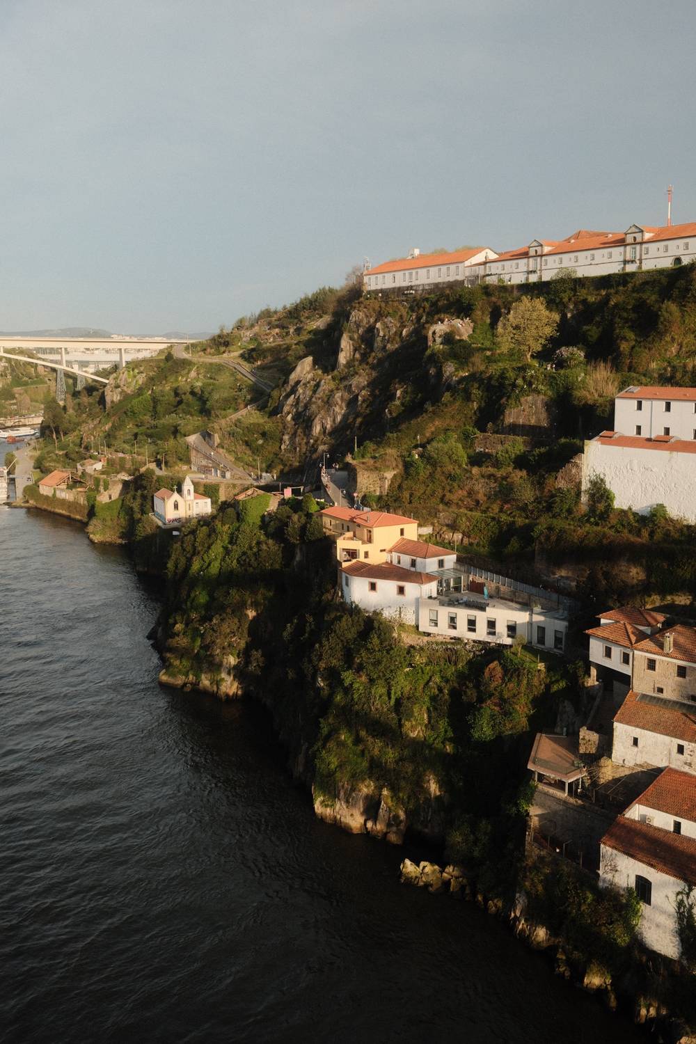 Aerial view of rocky cliffs along the Douro River in Porto, with white buildings and terracotta roofs nestled into the hillside and a large institutional building at the top.