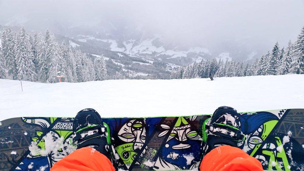 A point-of-view shot of feet strapped into a snowboard overlooking a snowy forested valley.