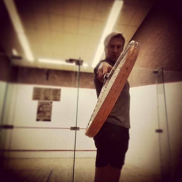 A man on an indoor squash court reaching out with his racket toward the camera.