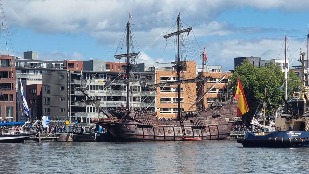 A wooden sailing ship moored in a harbour, with modern apartment buildings behind.