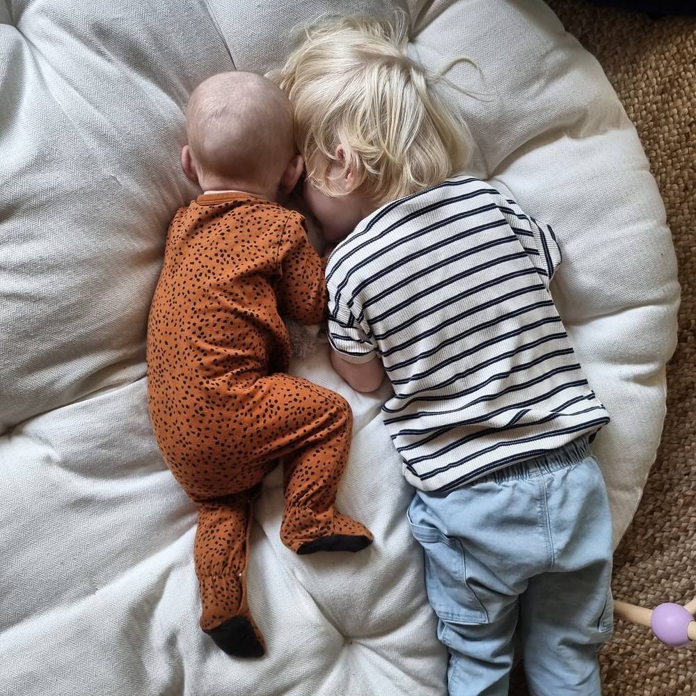 A baby in a leopard-print onesie and a toddler in a striped shirt lying head to head on a round cushion.